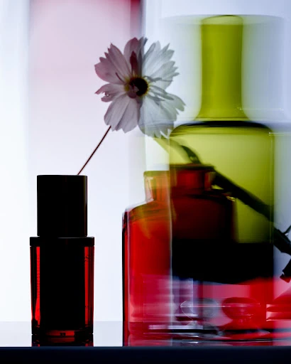 Still life shot of a single white daisy in a black vase, green glass and red glass bottles behind, against a white background.