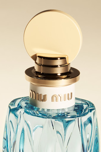Close-up of a Miu Miu perfume bottle with a light blue glass body, gold accents, and brand logo against a neutral backdrop.