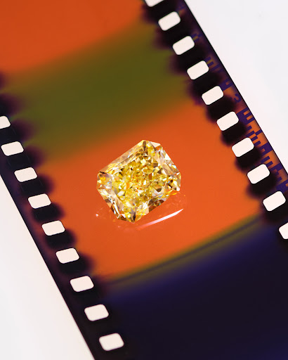 A bright yellow radiant-cut diamond sits on a light surface with a graphic film strip background with orange and black sections.