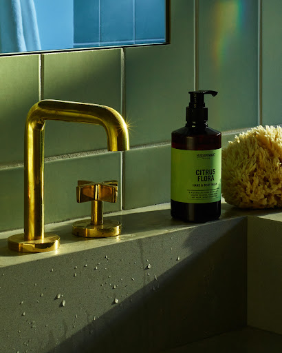 Modern bathroom still life featuring a gold faucet, soap dispenser, and sponge against a green tiled wall and concrete sink.