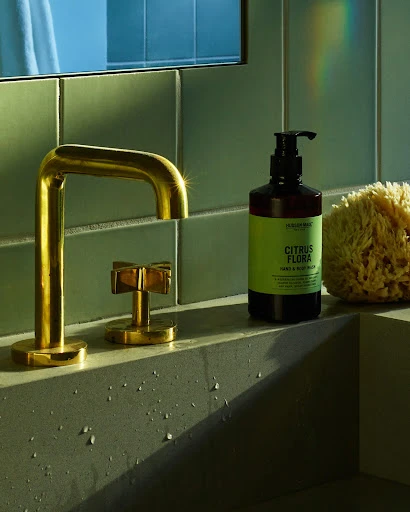 Modern bathroom still life featuring a gold faucet, soap dispenser, and sponge against a green tiled wall and concrete sink.