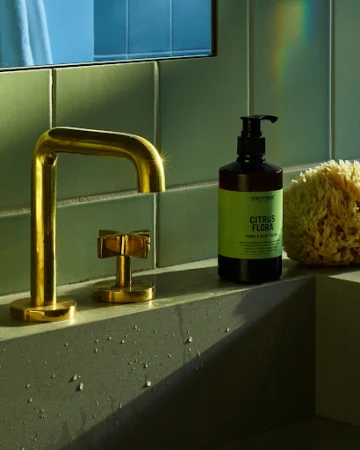Modern bathroom still life featuring a gold faucet, soap dispenser, and sponge against a green tiled wall and concrete sink.