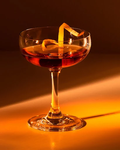 Crystal coupe glass containing an amber cocktail, garnished with an orange peel twist, against a dark, softly lit background.