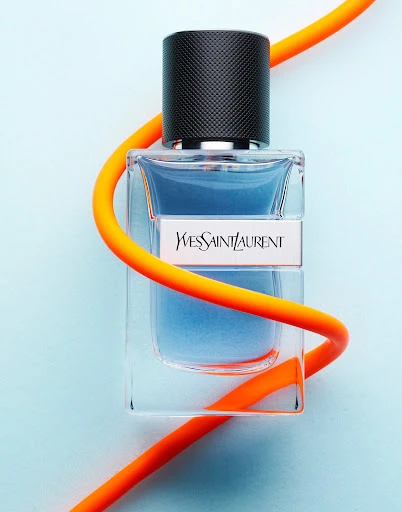 Studio shot of a YSL fragrance bottle with black cap, clear glass, and an orange cable, set against a soft blue backdrop.