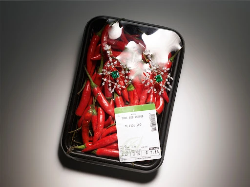 Close-up of red chili peppers arranged in a black plastic tray, adorned with silver and emerald jewelry, under studio lighting.