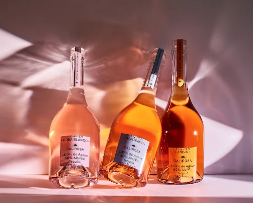 Three Calirosa tequila bottles sit on a shelf, featuring Rosa Blanco, Reposado, and Anejo varieties, casting shadows on the background.