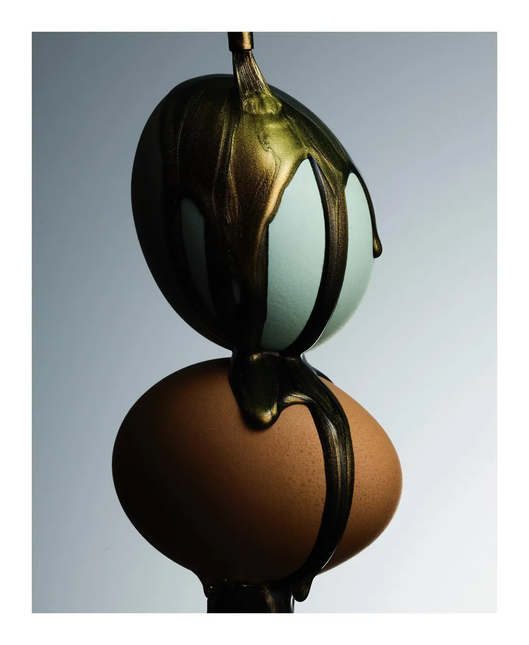 Two eggs stacked on top of each other with gold paint dripping down, studio shot. Conceptual food still life against gray gradient background.