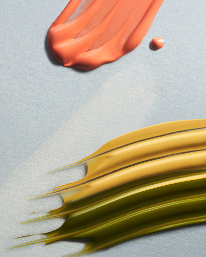 Abstract close-up of textured cosmetic smears in coral, yellow, and green on a light blue surface, casting soft shadows.