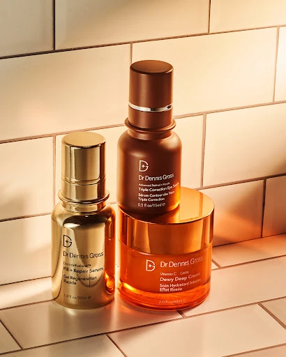 Dr. Dennis Gross skincare products including serum and cream containers in gold, amber, and brown hues against tile backdrop.