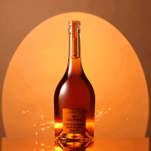 A bottle of Calirosa Tequila Anejo stands on a reflective surface against a warm, orange-lit background, illuminated by sparklers.