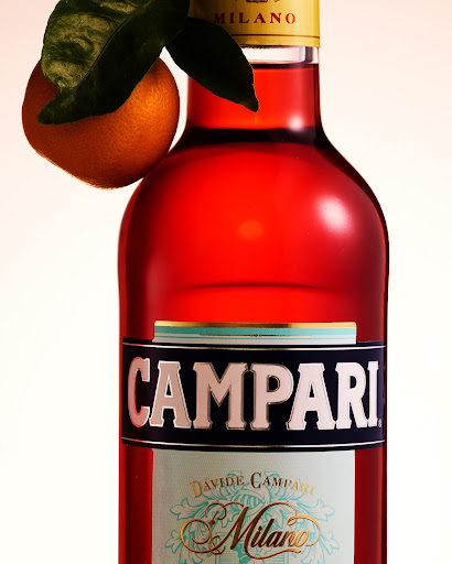 Close-up studio shot of a Campari Milano bottle with an orange and green leaves, set against a clean, evenly lit backdrop.