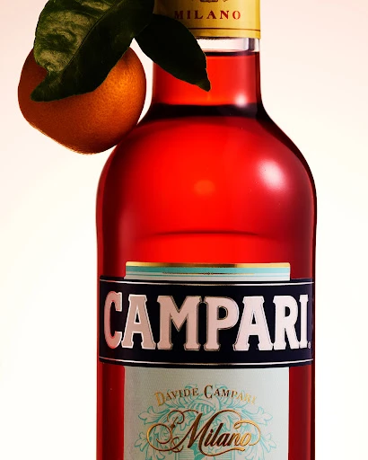 Close-up studio shot of a Campari Milano bottle with an orange and green leaves, set against a clean, evenly lit backdrop.