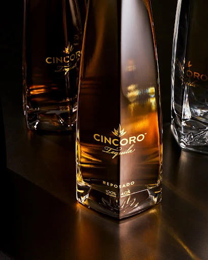 Three bottles of Cincoro Reposado Tequila on a dark surface in a studio shot, highlighting the bottles' angular design and golden liquor.