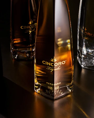 Three bottles of Cincoro Reposado Tequila on a dark surface in a studio shot, highlighting the bottles' angular design and golden liquor.
