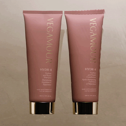 Two pink bottles of Vegamour HYDR-8 hydrate and repair shampoo and conditioner with gold lettering, on a neutral background.