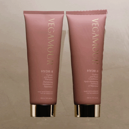 Two pink bottles of Vegamour HYDR-8 hydrate and repair shampoo and conditioner with gold lettering, on a neutral background.