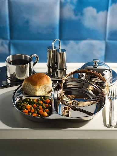 Shiny silver tray with food, gold jewelry, and drink presented on a white surface against a blue cloud-patterned background.
