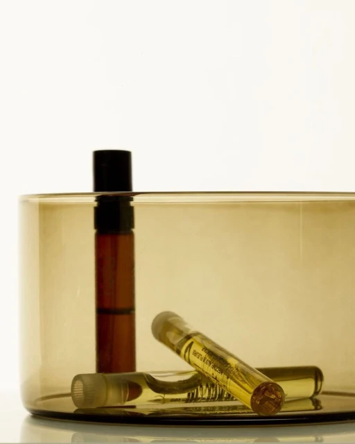 Two clear glass ampoules filled with yellow liquid and a brown cosmetic bottle rest inside a brown glass container with a white backdrop.