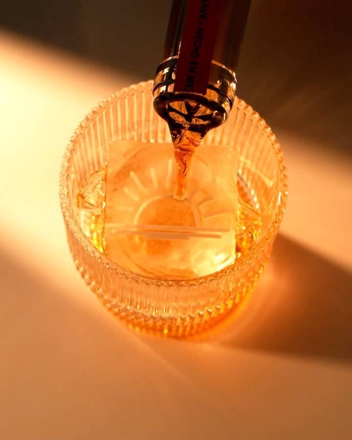 Whiskey is poured from a bottle into a glass with a large ice cube, creating a visually striking and refreshing drink presentation.