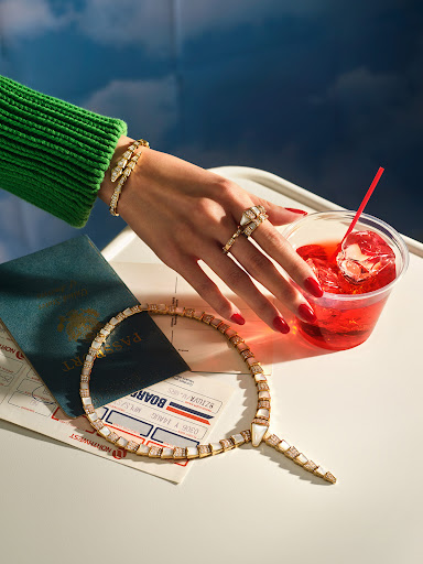 Hand with rings and bracelet next to a drink with a straw, passport, airplane ticket, and necklace on a white table with a blue background.