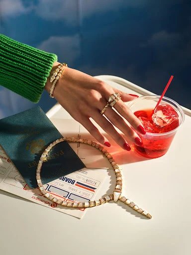 Hand with rings and bracelet next to a drink with a straw, passport, airplane ticket, and necklace on a white table with a blue background.