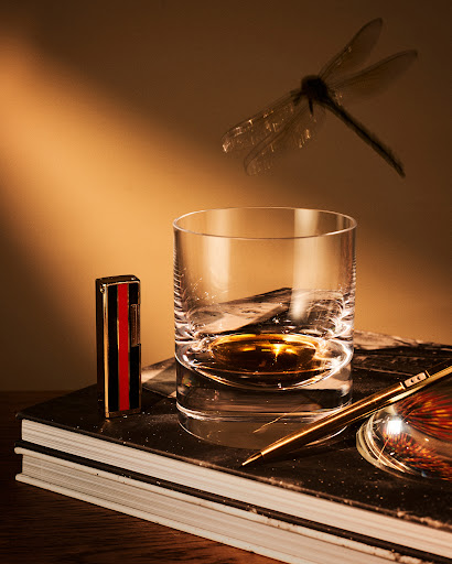 Whiskey glass with liquid and vintage lighter on stack of books, with a pen, fishing lure, and dragonfly in a warm, golden light.