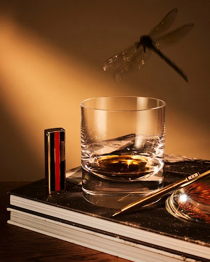 Whiskey glass with liquid and vintage lighter on stack of books, with a pen, fishing lure, and dragonfly in a warm, golden light.