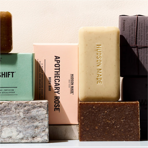 Close-up shot of stacked Hudson Made body bars in varying scents: Apothecary Rose, Shift, and a brown bar, staged in a studio setting.