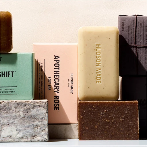 Close-up shot of stacked Hudson Made body bars in varying scents: Apothecary Rose, Shift, and a brown bar, staged in a studio setting.