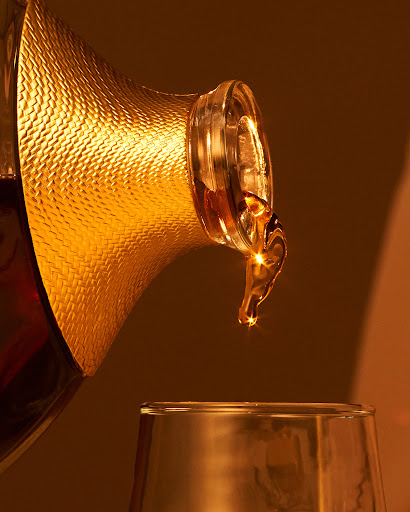 Golden liquid is poured from a patterned decanter into a clear glass, showcasing an amber color and dynamic pouring motion.
