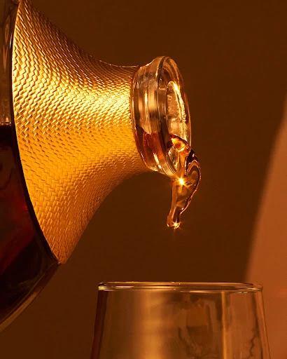 Golden liquid is poured from a patterned decanter into a clear glass, showcasing an amber color and dynamic pouring motion.
