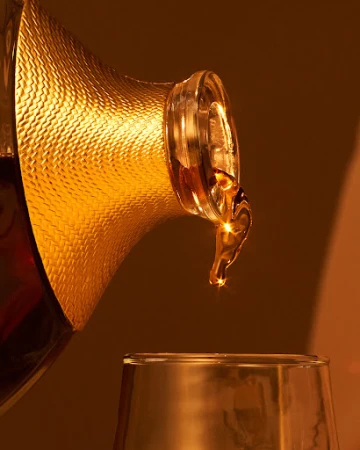 Golden liquid is poured from a patterned decanter into a clear glass, showcasing an amber color and dynamic pouring motion.