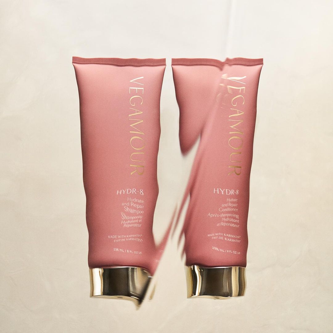 Two Vegamour HYDR-8 hair product tubes sit side-by-side on a reflective surface, featuring pink packaging with gold lettering.