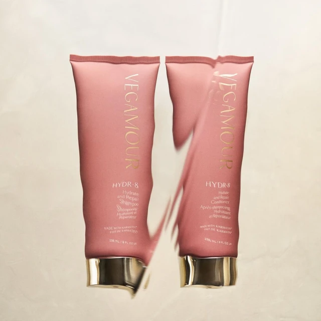 Two Vegamour HYDR-8 hair product tubes sit side-by-side on a reflective surface, featuring pink packaging with gold lettering.