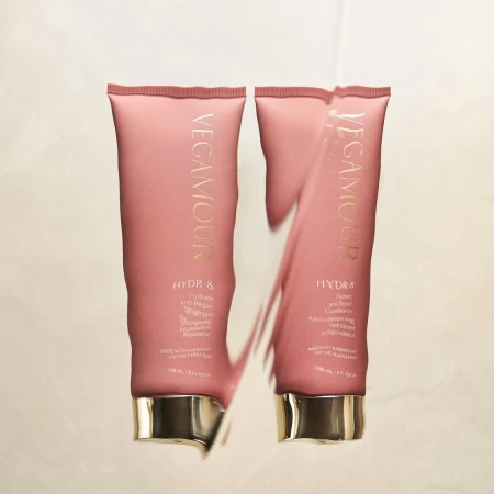 Two Vegamour HYDR-8 hair product tubes sit side-by-side on a reflective surface, featuring pink packaging with gold lettering.