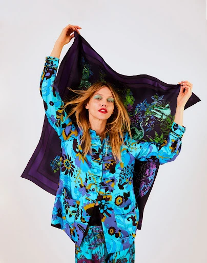 Adult woman with floral shirt and pants holding patterned scarf up against a white background in a colorful fashion pose.