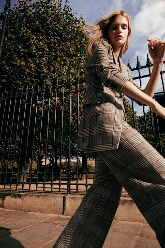 A blonde woman in a plaid suit walks outside next to a fence and green trees under a cloudy sky in a fashion editorial image.