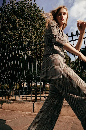 A blonde woman in a plaid suit walks outside next to a fence and green trees under a cloudy sky in a fashion editorial image.