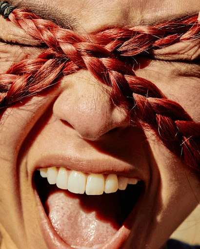 Close up of a screaming adult woman's face with red braids obscuring her eyes, captured in bright, natural light.