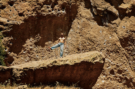 Adult male standing on a rock formation in blue jeans, posing against a background of large rock formations with sparse vegetation.