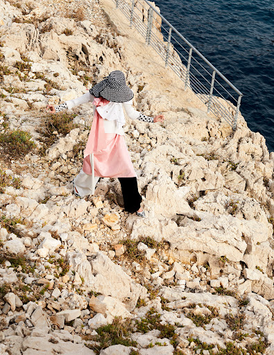 A woman in stylish clothing stands on a rocky cliff overlooking the sea. She is wearing a checkered hat and fashionable outfit.