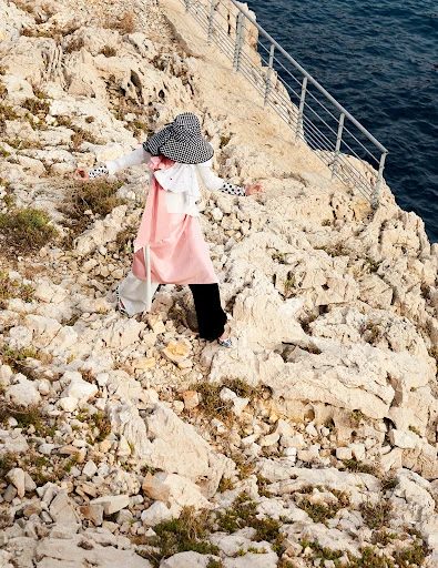 A woman in stylish clothing stands on a rocky cliff overlooking the sea. She is wearing a checkered hat and fashionable outfit.