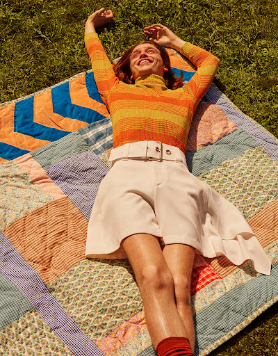 Adult laying on a quilt in the grass wearing orange striped turtleneck, white shorts, and orange socks with arms raised outdoors