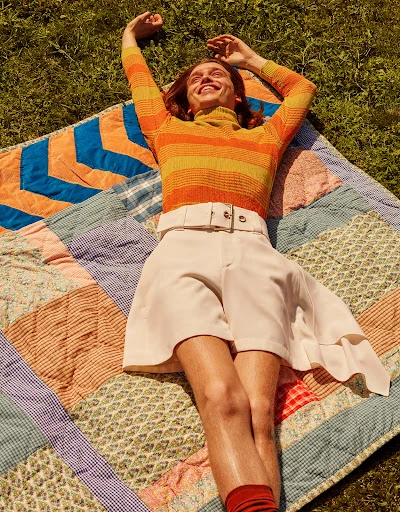 Adult laying on a quilt in the grass wearing orange striped turtleneck, white shorts, and orange socks with arms raised outdoors