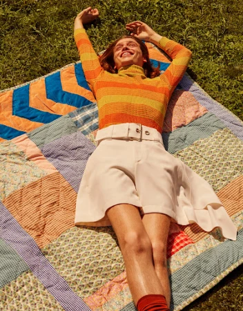 Adult laying on a quilt in the grass wearing orange striped turtleneck, white shorts, and orange socks with arms raised outdoors