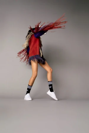 Model wearing fringed garment and black socks with white sneakers dancing against grey studio background