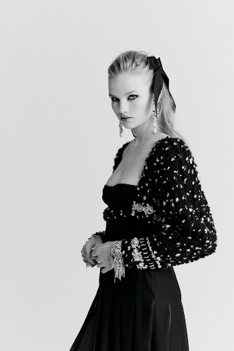 Black and white portrait of an adult woman in elegant black evening wear with decorative jacket, jewelry, and a large hair bow.