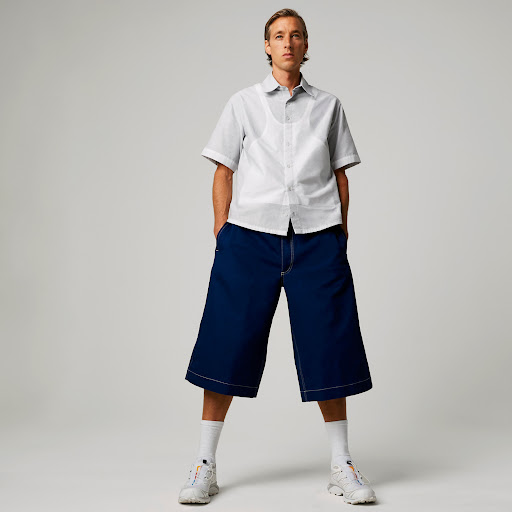 Full-length shot of a young adult man in an open-collar shirt, wide-leg cropped pants, and white sneakers posing in a photo studio.