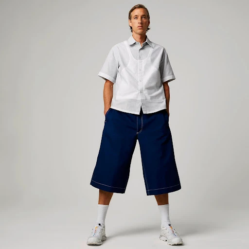 Full-length shot of a young adult man in an open-collar shirt, wide-leg cropped pants, and white sneakers posing in a photo studio.