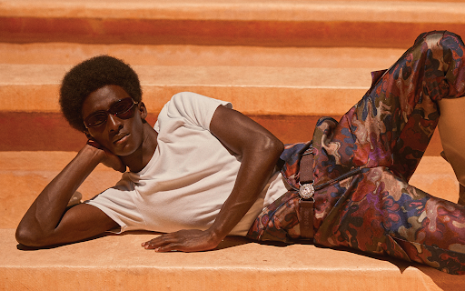 Man with afro and sunglasses reclines on orange steps wearing a white t-shirt and patterned trousers in a fashion pose.